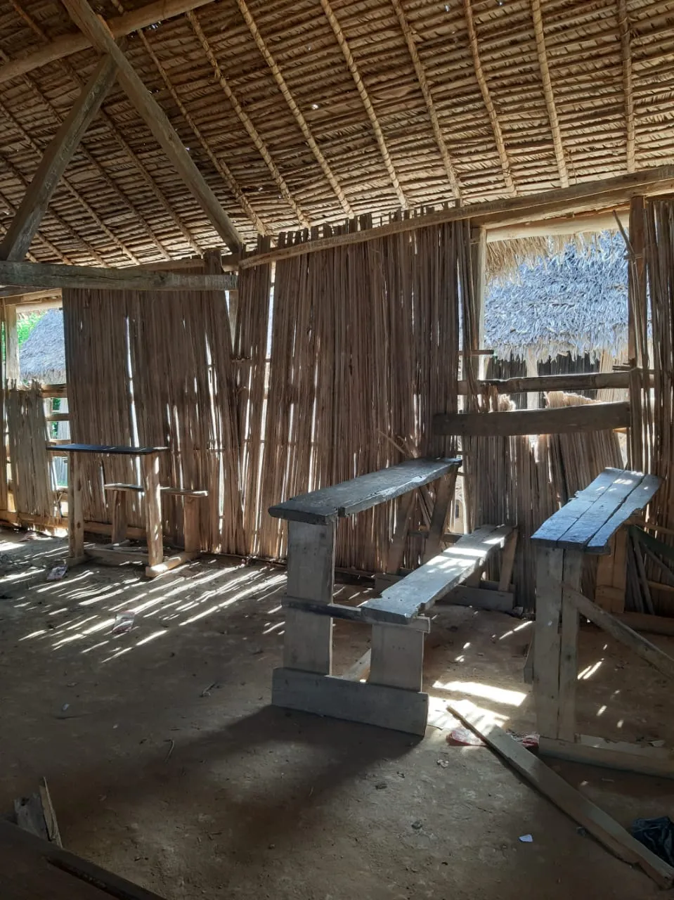 Une salle de classe aux murs de lattes sous un toit de chaume, ses tables-bancs usés